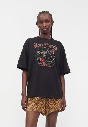 Vrouw met gevlochten haar, die een oversized zwart Von Dutch T-shirt met panterafbeelding en korte broek met luipaardprint draagt, staand tegen een effen achtergrond.