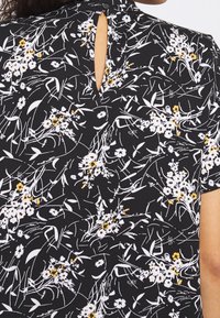 Blouse noire à fleurs avec des fleurs blanches et jaunes, tissu doux, encolure ronde, manches courtes et un détail goutte avec un bouton au dos.