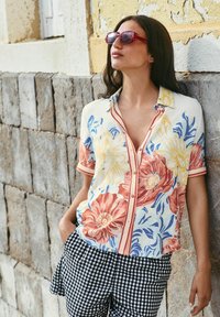 Bloemige korte mouwen shirt in lichte stof, met grote gele en rode bloemen. Gepaard met zwart-witte geblokte broek. Rode zonnebril.