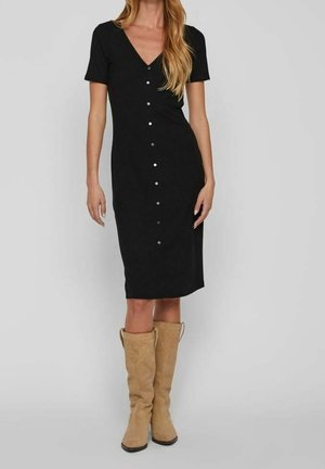 Zwarte geribbelde midi-jurk met korte mouwen, V-hals en knoopdetails aan de voorkant. Gepaard met tan kniehoge suède laarzen.