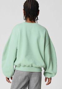Felpa oversize verde menta con spalle basse e polsini a costine. Tessuto morbido, vestibilità ampia e orlo elastico. Indossata con pantaloni grigi.