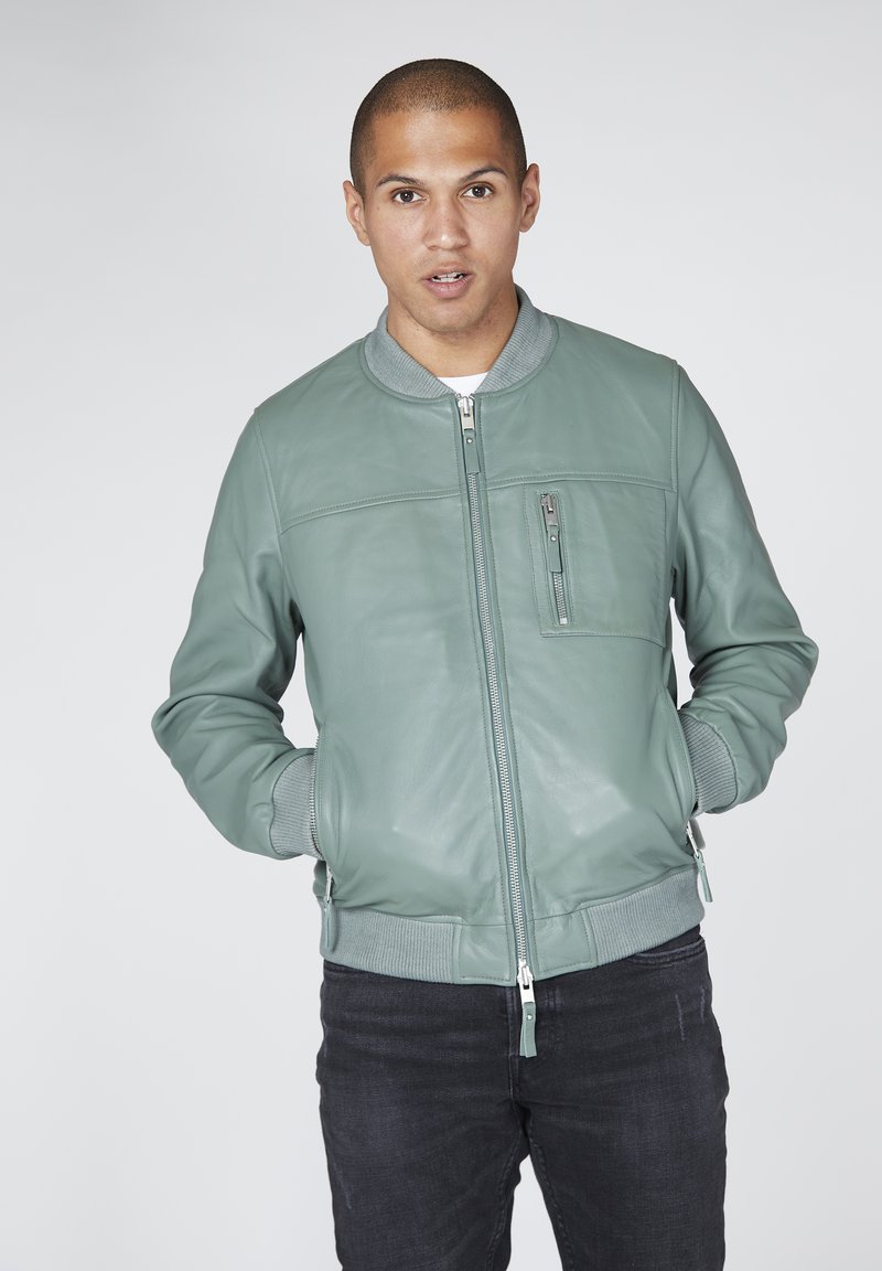 Zelená kožená bomber bunda s žebrovaným šedým límečkem a manžetami, se zipovou náprsní kapsou a bočními kapsami. Nošena s tmavými džíny.