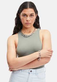 Grøn ærmeløs bodysuit med en glat tekstur, sølv kæderekæde og armbånd, kvinde med armene korslagte, lyse denim-shorts.