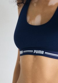 Navy tkanina sportski grudnjak s izrezom u obliku šalice i PUMA logom na elastičnom remenu; ima glatku teksturu i prilagođeni dizajn.