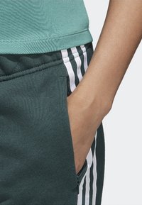 Green athletic top paired with dark green pants featuring white vertical stripes. Close-up shows a hand in the pocket and fabric texture.