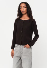 Suéter de malha castanho-escuro com decote redondo, mangas longas, fecho com botões à frente e texturização subtil. Combinado com jeans cinza-claro.