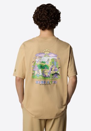 Osoba z kręconymi włosami nosi beżowy t-shirt z kolorową sceną z kreskówki przedstawiającą słońce, góry, kwiaty, zwierzęta i tekst "Explorers Gather."