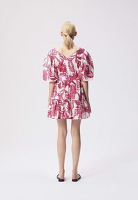 Woman with blonde hair in a ponytail wearing a short white dress with pink floral print and puff sleeves, standing on a plain white background.