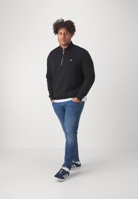 Felpa nera con zip e logo giallo, jeans blu e scarpe da ginnastica blu scuro. Il tessuto sembra morbido con una texture liscia.