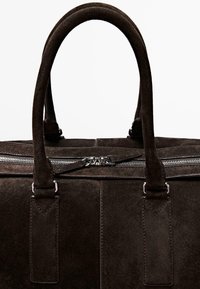 Tmavě hnědá semišová taška se dvěma pevnými držadly, stříbrným zipem a texturovanými detaily. Plochý design bez viditelného kovového příslušenství.