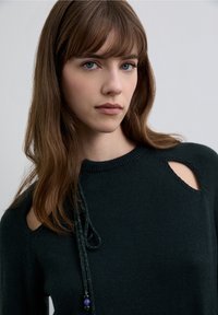 Tmavě zelený pletený svetr s vykrojenými rameny, kulatým výstřihem a ozdobným šňůrkovým detailem s korálky. Hladká textura a přiléhavý střih.
