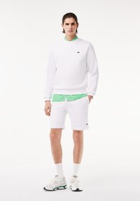 Weißer Kapuzenpullover und Shorts, darunter ein grünes Hemd mit Kragen, das ein kleines Logo auf der linken Brust trägt, kombiniert mit weißen Sportschuhen.