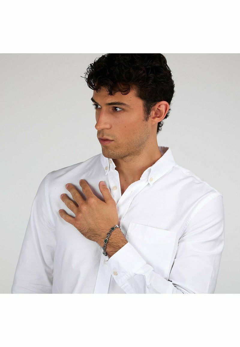 Witte button-up shirt met een borstzak, gedragen door een persoon die hun borst aanraakt. Ze dragen een zilveren kettingarmband om één pols.