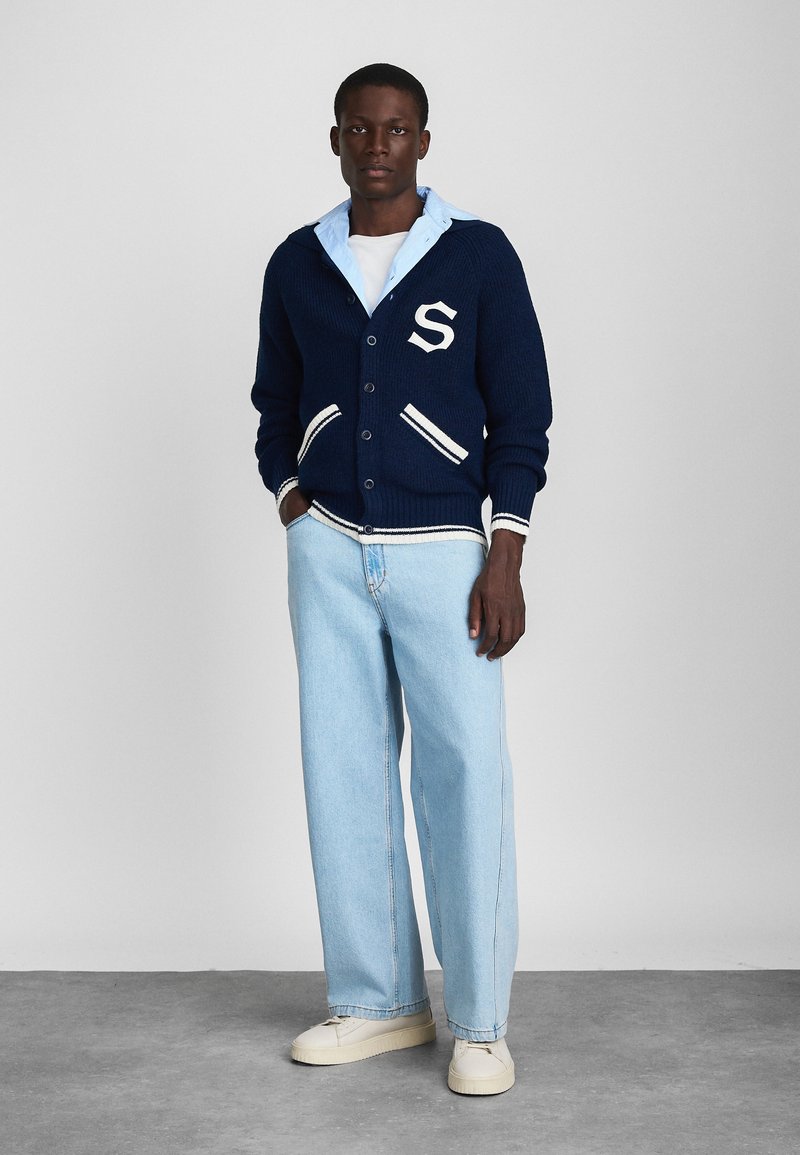 Marineblauwe gebreide cardigan met witte accenten en de letter "S", gedragen over een lichtblauw overhemd met kraag en gecombineerd met lichtblauwe jeans en crèmekleurige sneakers.