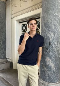 Tânăr îmbrăcat într-un tricou polo albastru închis și pantaloni bej se sprijină de o coloană mare de piatră lângă o ușă ornamentată, privindu-se gânditor în lateral.