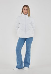 Giacca imbottita bianca con colletto alto e chiusura frontale a zip. Presenta un motivo a trapuntatura geometrica. Abbinata a jeans a zampa d'elefante di colore blu chiaro e scarpe bianche.