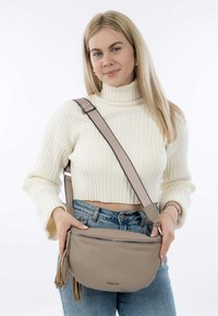 Beige Leder-Umhängetasche mit Reißverschluss, strukturierten Oberflächen und Quastendetail. Getragen mit einem hellen Pullover und blauen Jeans.