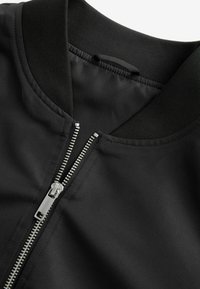 Cerca de una chaqueta bomber negra, con una tela suave, cuello acanalado y cremallera metálica con un tirador plateado, todo sobre un fondo mate.
