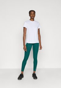 Balti trumpomis rankovėmis marškinėliai derinami su žaliomis aptemptomis tamprėmis su užtrauktukų detalėmis, papildytos juodais sportiniais bateliais.