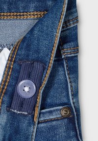 Denim tkanina s plavim tonovima, sa svijetlosmešavim šavovima. Uključuje tamnu dugmad s prozirnim dugmetom i metalnim dugmetom na pritiskanje.