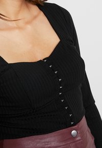 Mustassa ribatulla paidalla on neliönmuotoinen pääntie, pitkät hihat ja hopeiset nappikiinnitykset edessä. Rakenne- ja tekstuuri lisäävät syvyyttä.