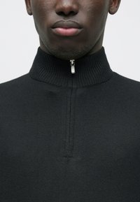 Schwarzer Strickpullover mit hohem Kragen und kurzem Reißverschluss am Hals. Glatte Textur mit figurbetontem Schnitt.