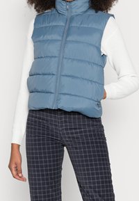 Modrá prošívaná vesta s vysokým límcem, zipem na přední straně a prošívaným vzorem. Nošena přes bílý top s dlouhým rukávem a doplněna kostkovanými šedými kalhotami.