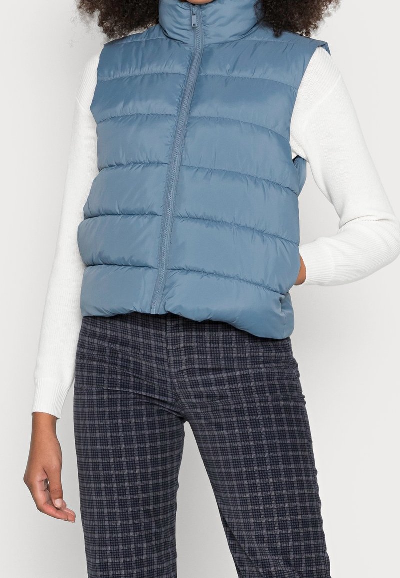 Modrá prošívaná vesta s vysokým límcem, zipem na přední straně a prošívaným vzorem. Nošena přes bílý top s dlouhým rukávem a doplněna kostkovanými šedými kalhotami.