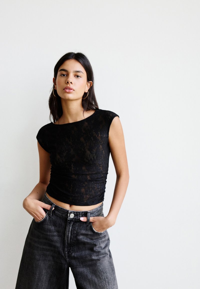Schwarzes Spitzen-Crop-Top mit kurzen Ärmeln, das einen taillierten Oberkörper und geraffte Textur aufweist, kombiniert mit hochgeschnittenen grauen Jeans.