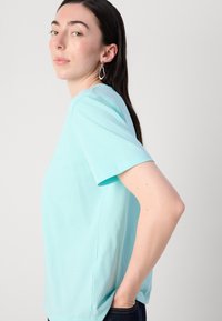 Svetlo modré bavlnené tričko s krátkymi rukávmi, okrúhlym výstrihom a voľným strihom. Model stojí bokom, s rukami v vreckách. Hladká textura.