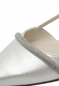 Stříbrná metalická plochá bota s špičatou špičkou, která má texturovaný pásek zdobený malými kulatými lesklými ozdobami.