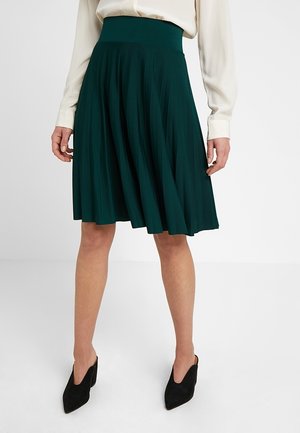 Mujer con una falda plisada verde oscuro hasta la rodilla, blusa de manga larga color crema y zapatos de tacón negros con punta puntiaguda, de pie sobre un suelo blanco.