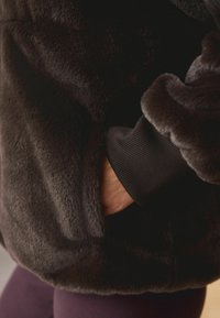 Jachetă din blană sintetică de culoare gri închis, cu o textură moale, având un manșon ribat de culoare neagră și un buzunar lateral integrat în design.