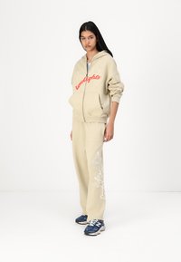 Beige hoodie with red text, zippered front, and front pockets. Matching beige sweatpants with white graphic design on the side. Blue sneakers.