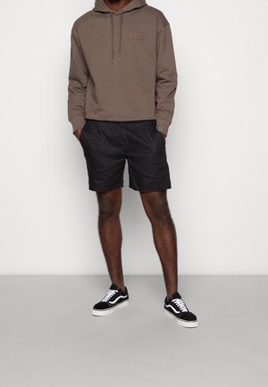 Sudadera marrón con capucha y un pequeño parche de logo, combinada con shorts negros y zapatillas negras con acentos blancos. Conjunto informal expuesto.