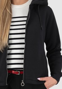 Felpa nera con zip e texture morbida, dotata di cappuccio con cordoncino e tasche laterali, indossata sopra un maglione a righe bianche e nere.