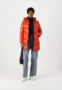 Donsjas in glanzend rood, met een capuchon, ritssluiting en zijzakken. Gecombineerd met lichtgewassen, wijde jeans en witte sneakers.