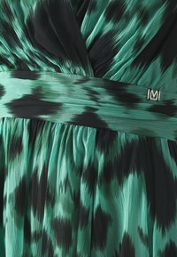 Robe sans manches verte et noire avec une texture plissée, présentant un design superposé et un accent en argent en forme de "M" sur la taille.
