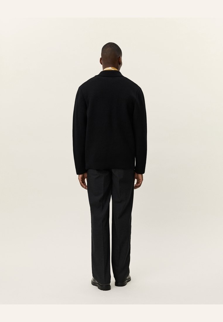 Un homme est debout de dos, portant une veste noire, un pantalon noir, des chaussures noires et un col de chemise clair visible au niveau du cou.