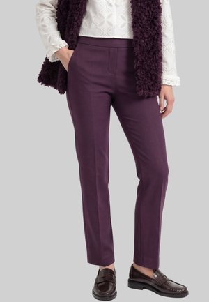 Woman wearing straight-fit purple trousers, white lace blouse, dark purple textured vest, and dark brown loafers, standing with one hand in pocket.