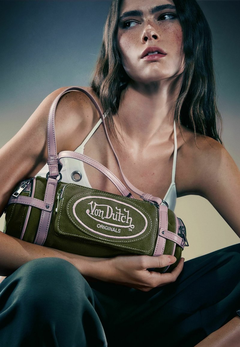 Giovane donna con capelli lunghi che tiene una borsa a tracolla Von Dutch Originals verde e rosa, indossa una canotta bianca e pantaloni scuri su uno sfondo semplice.