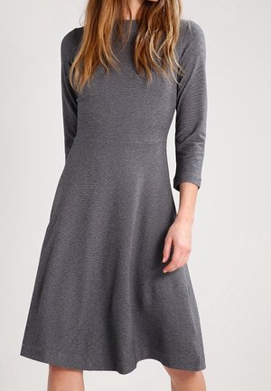 Kobieta ubrana w szarą, teksturowaną sukienkę do kolan z rękawami do połowy przedramion, stojąca na tle jednolitego, jasnego tła.