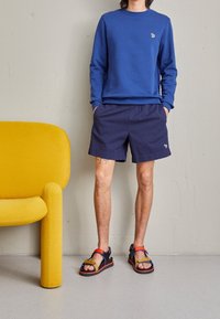 Sudadera azul, pantalones cortos azul marino con bordado y sandalias multicolores con correas. Silla amarilla en el fondo, textura suave, diseño moderno.