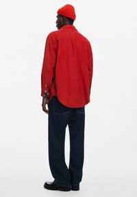 Rød langærmet skjorte med en rygfaltung, parret med mørkeblå brede jeans og sorte støvler. Skjorten har subtile mønstre og knaplukninger ved håndleddene.
