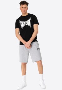 Čierne bavlnené tričko s bielym logom "TAPOUT"; šedé šortky s logom na ľavom stehne; biele tenisky; póza v stoji na neutrálnom pozadí.