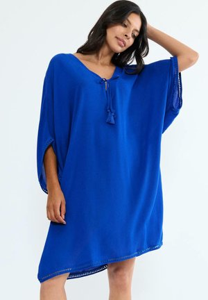 Frau trägt ein locker sitzendes königsblaues Tunika-Kleid mit weiten Ärmeln und Quastenverschluss am Ausschnitt, posiert mit einem erhobenen Arm.