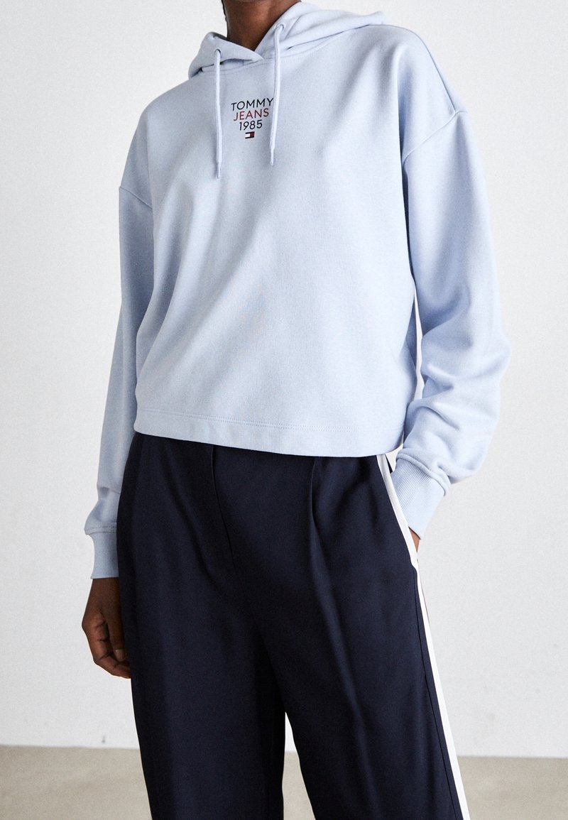Personne portant un sweat à capuche court bleu clair avec le texte « Tommy Jeans 1985 », associé à un pantalon bleu marine avec des bandes blanches sur les côtés.
