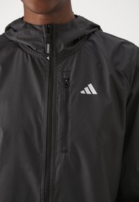 Czarna kurtka biegowa z kapturem, z pełnym zamkiem błyskawicznym, przednią kieszenią oraz odblaskowym logo Adidas na piersiach.