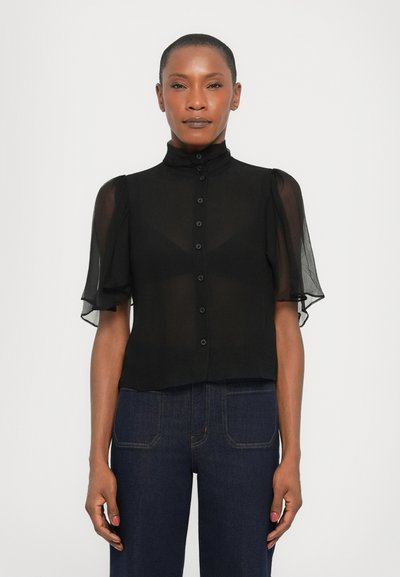 Blouse noire transparente avec des manches bouffantes courtes, un devant à boutons, un col haut et un design légèrement court. Portée avec un pantalon en denim noir.