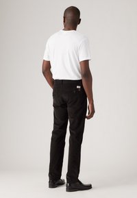 Pantalones de pana negros con un corte recto, que cuentan con cinco bolsillos, una etiqueta en la parte trasera y una superficie acanalada texturizada. Llevados con una camiseta blanca.